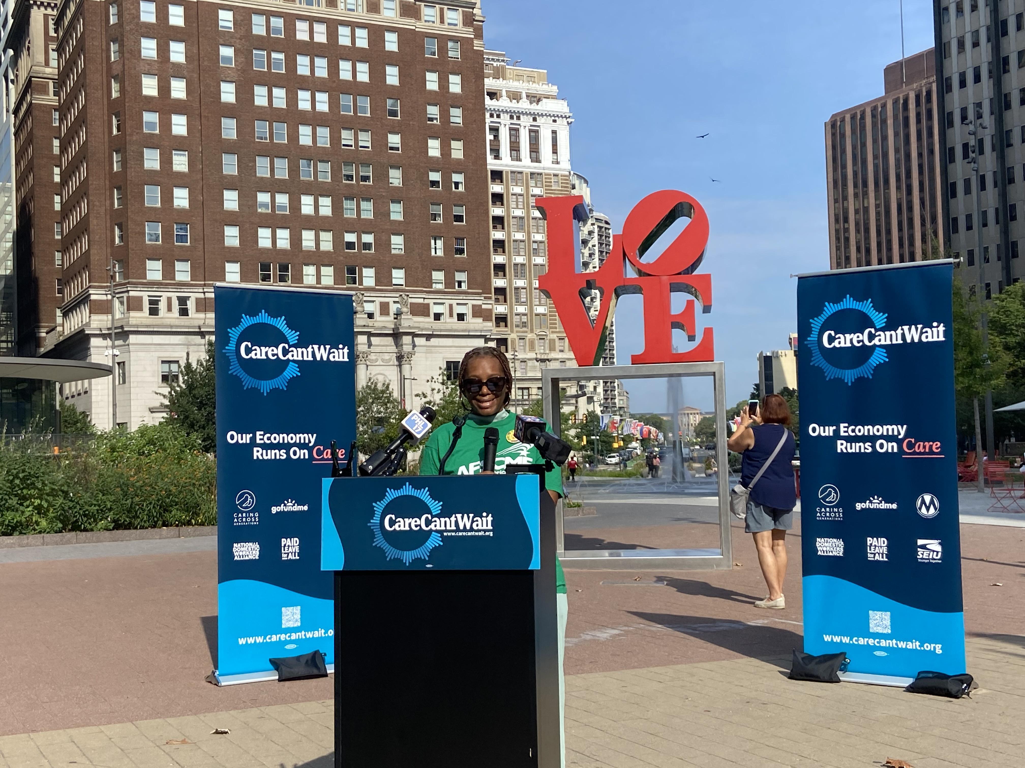 Woman in green AFSCME shirt speaking at podium in front of Care Cant Wait banners. 